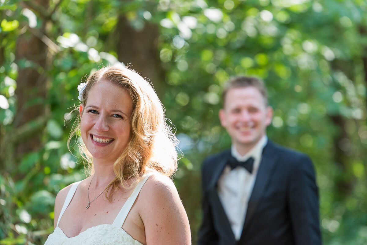 Ein Hochzeitspaar. Die Braut steht im weißen Kleid im Vordergrund. Der Bräutigam steht etwas weiter hinten in der Unschärfe.