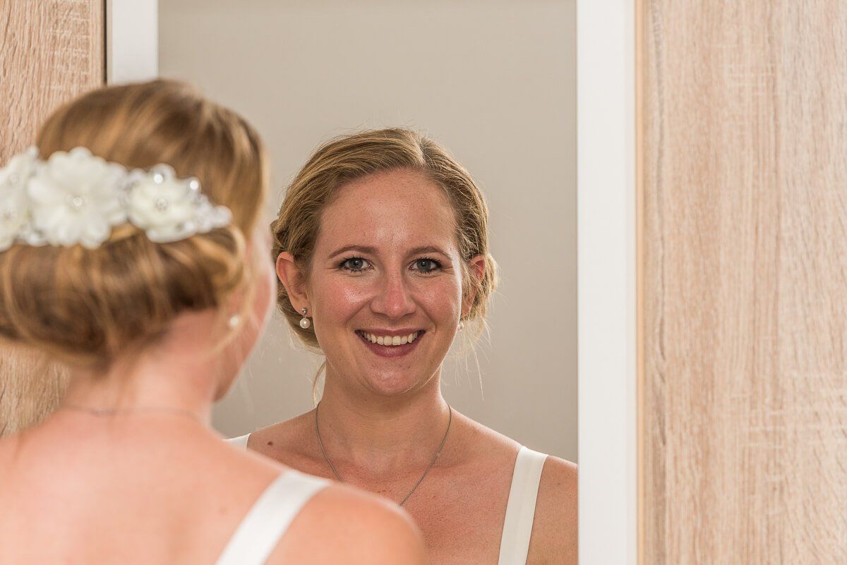 Eine Braut im weißen Kleid mit hochgesteckten Haaren sieht von hinten fotografiert durch einen Wandspiegel lächelnd in die Kamera.
