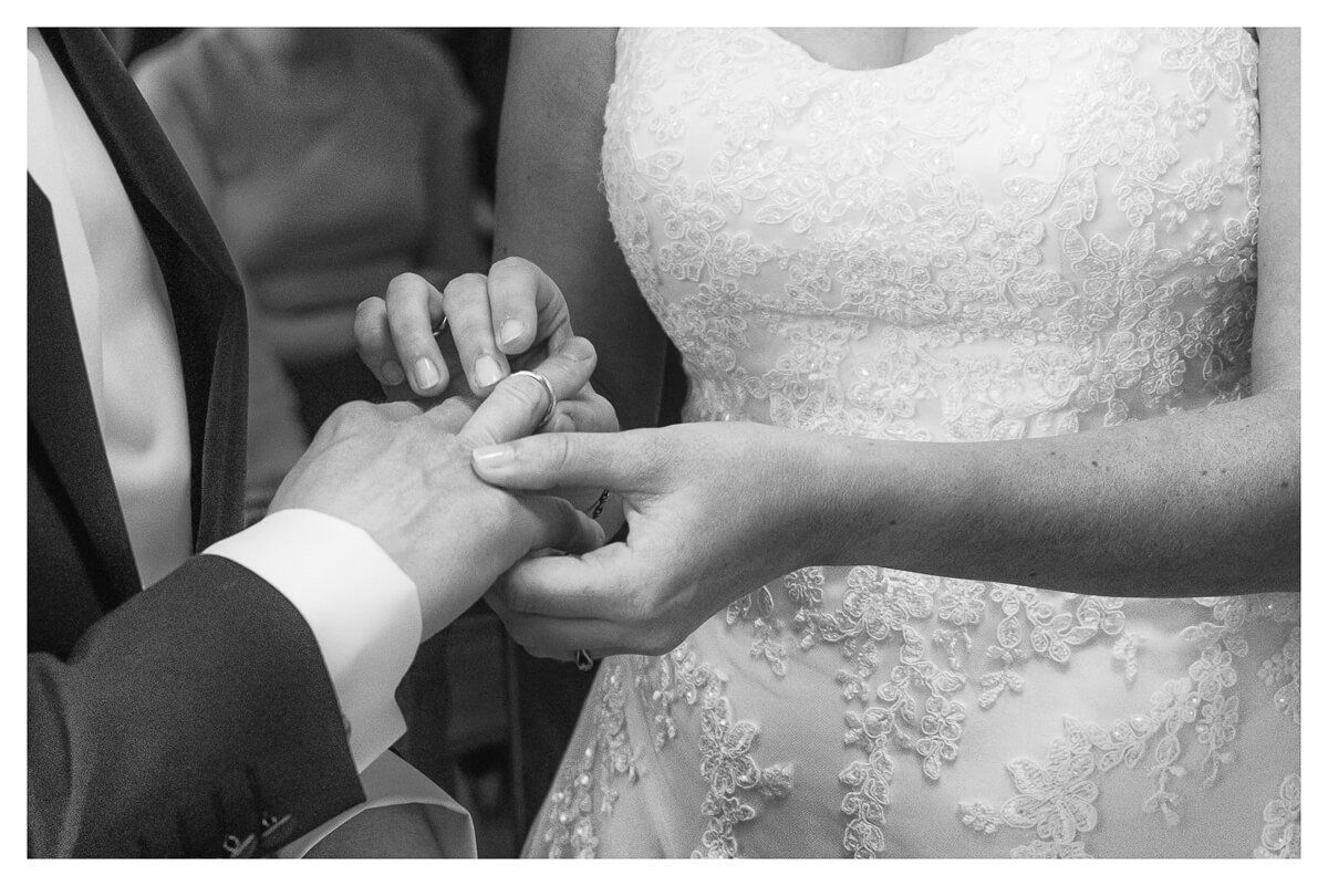 Eine Braut im weißen Kleid steckt ihrem Bräutigam den Ring an den Finger.