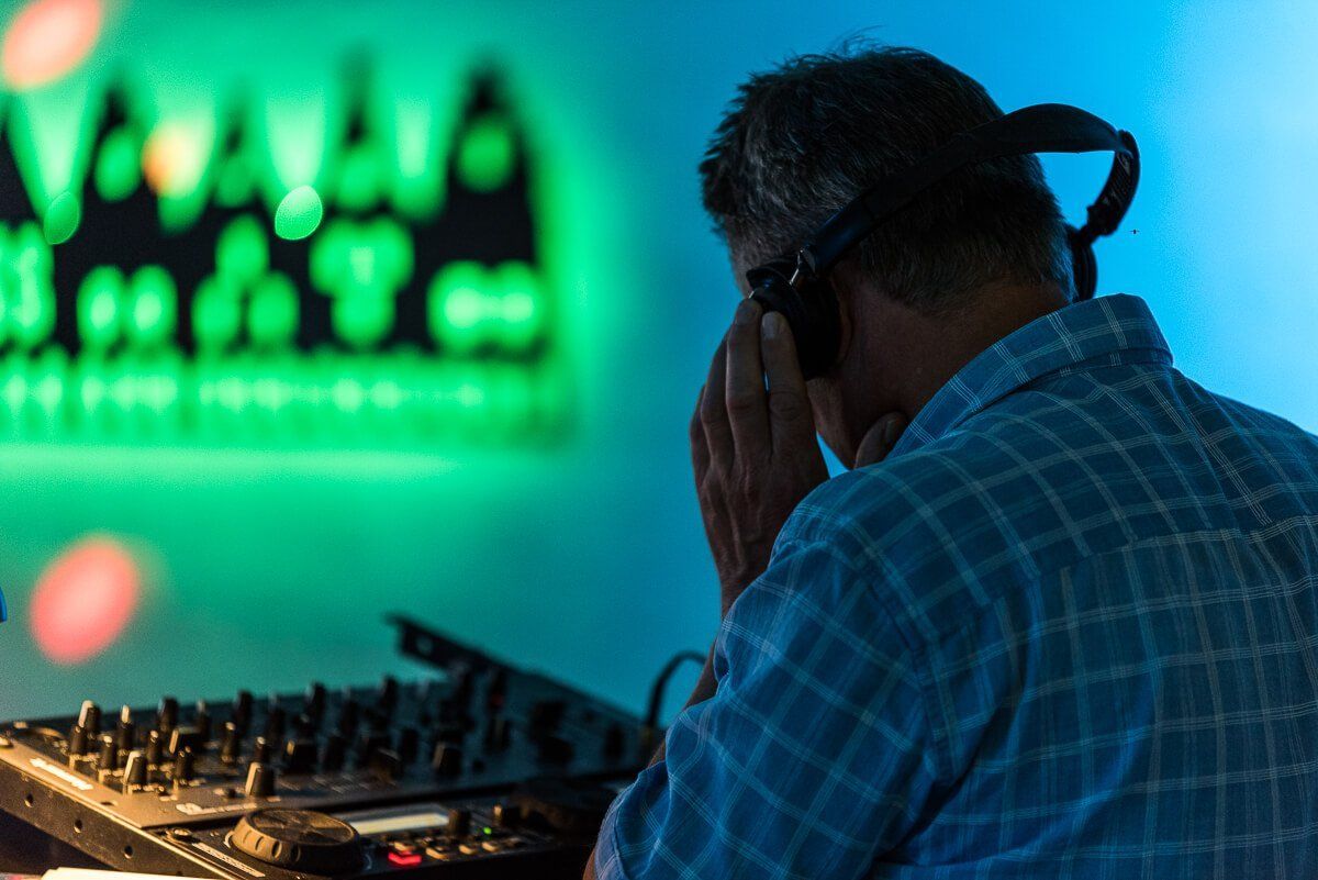 Ein DJ schräg seitlich von hinten fotografiert. Er hat seine linke Hand am Kopfhörer.