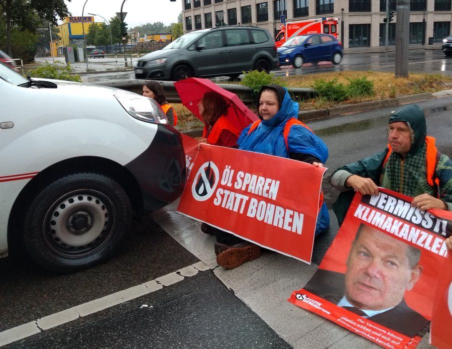 Straßenblockade, Regen, direkt vor einem weißen Auto, blockierende. Ihre Schilder: Öls sparen statt bohren. Vermisst: Klimakranzler. Logo der #LetztenGeneration