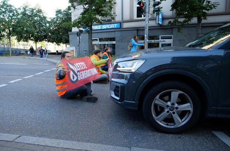 Straßenblockade der Letzten Generation in Stuttgart. Seitlich aufgenommen: Ein größeres Auto steht dicht vor dem vordersten Aktivisten. Quelle: https://letztegeneration.de/presse/pressebilder/ von Achim Hegeler
