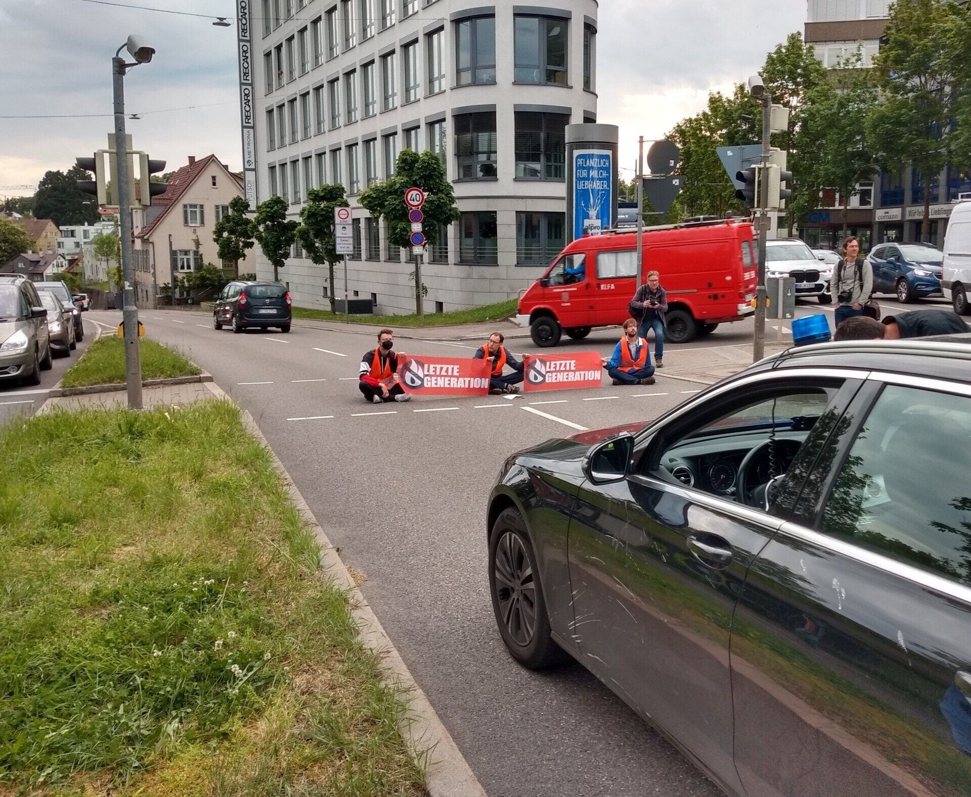 Drei Menschen der letzten Generation sitzen auf der Straße und blockieren zwei Fahrbahnen, zwischen ihnen zwei Banner mit Namen und Logo der Initiative, im Vordergrund ein Stück eines dunklen PKW (mit Blaulicht auf dem Dach), im Hintergrund fahren ein PKW und ein kleiner Feuerwehrwagen von einer Nebenstraße, die hinter der Blockadestelle auf die Fahrbahn führt, hinter der Blockade auf die Straße,