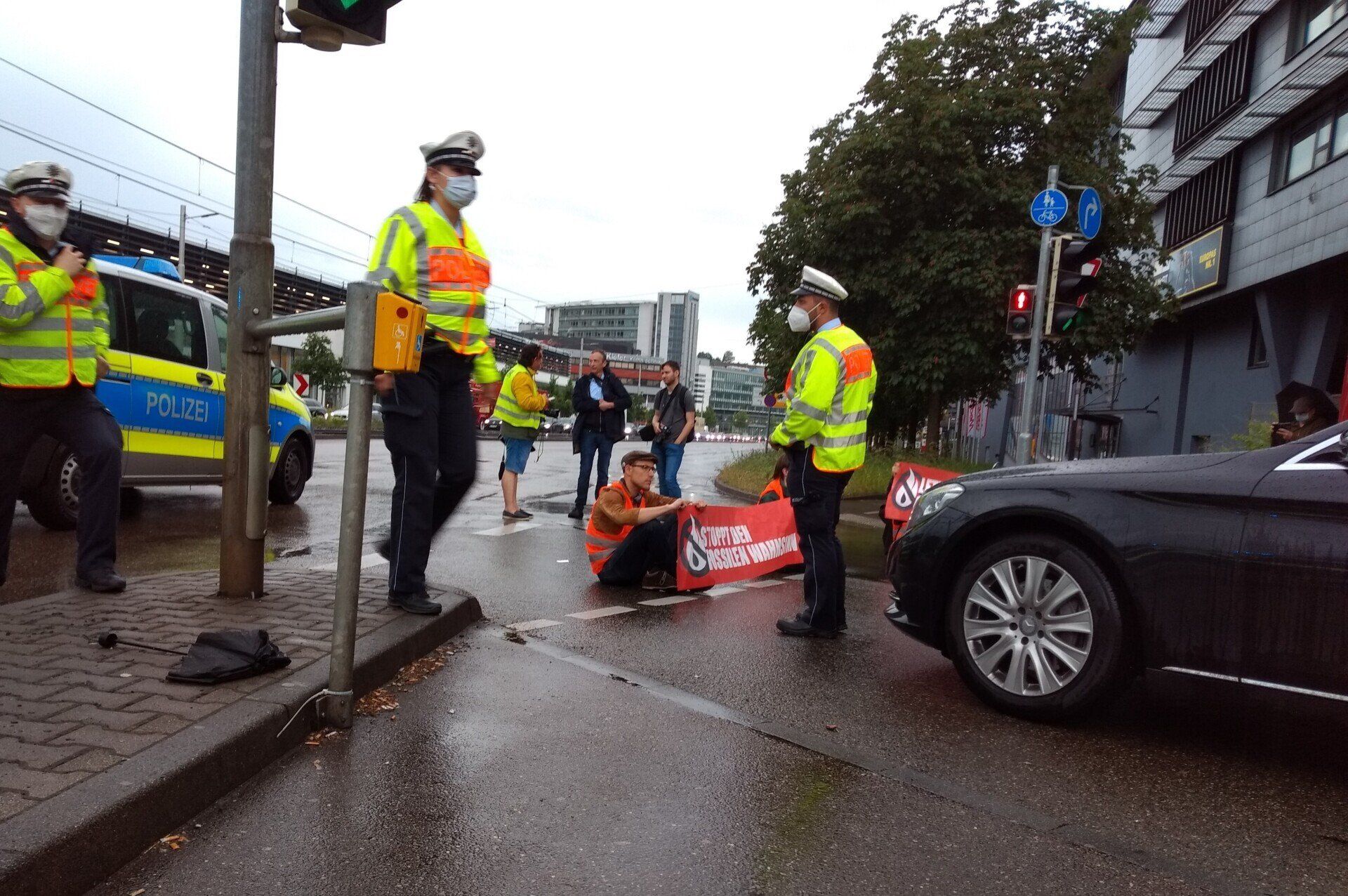 Nasse Straße, rechts ein Stück eines dunklen PKW, im Hintergrund ein Polizeiwagen, ein Polizist steht vor der Blockade von drei Menschen mit Bannern, zwei gehen, im Hintergrund ein Polizeiwagen