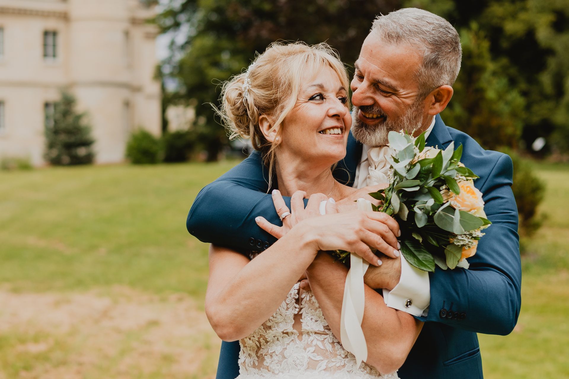 Couple de mariés enlacés devant un château, la mariée tenant un bouquet de fleurs jaunes et vertes