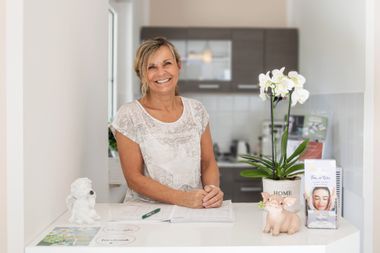 Ilka Schreiter Bild der Inhaberin Ilka Schreiter. Sie steht an einem Tresen, daneben sind Orchideen. Ein Engel und ein Sparschweinchen stehen auch auf dem Tresen.