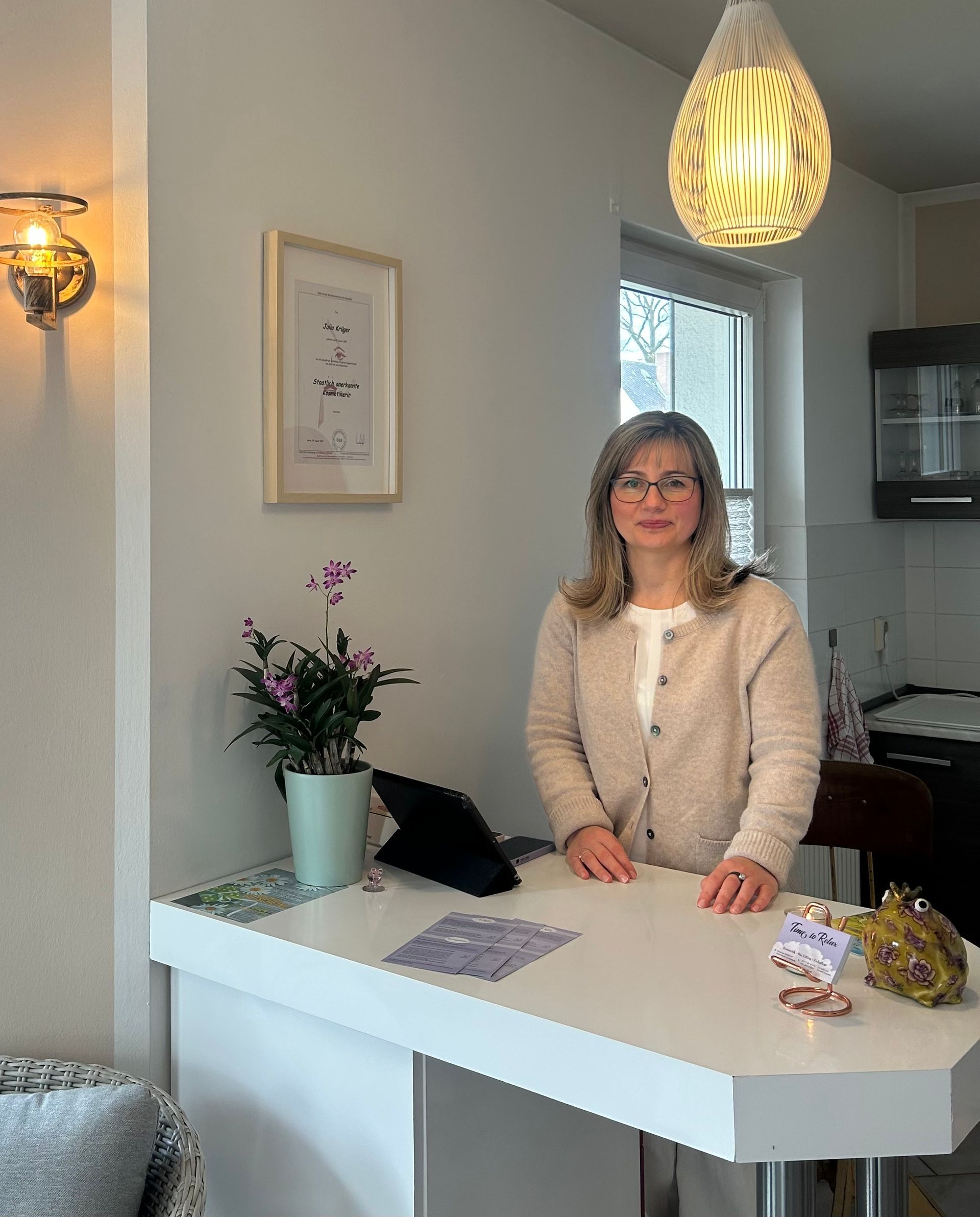 Julia Krüger Bild der Inhaberin Julia Krüger. Sie steht an einem Tresen, daneben sind Orchideen. Ein Engel und ein Sparschweinchen stehen auch auf dem Tresen.
