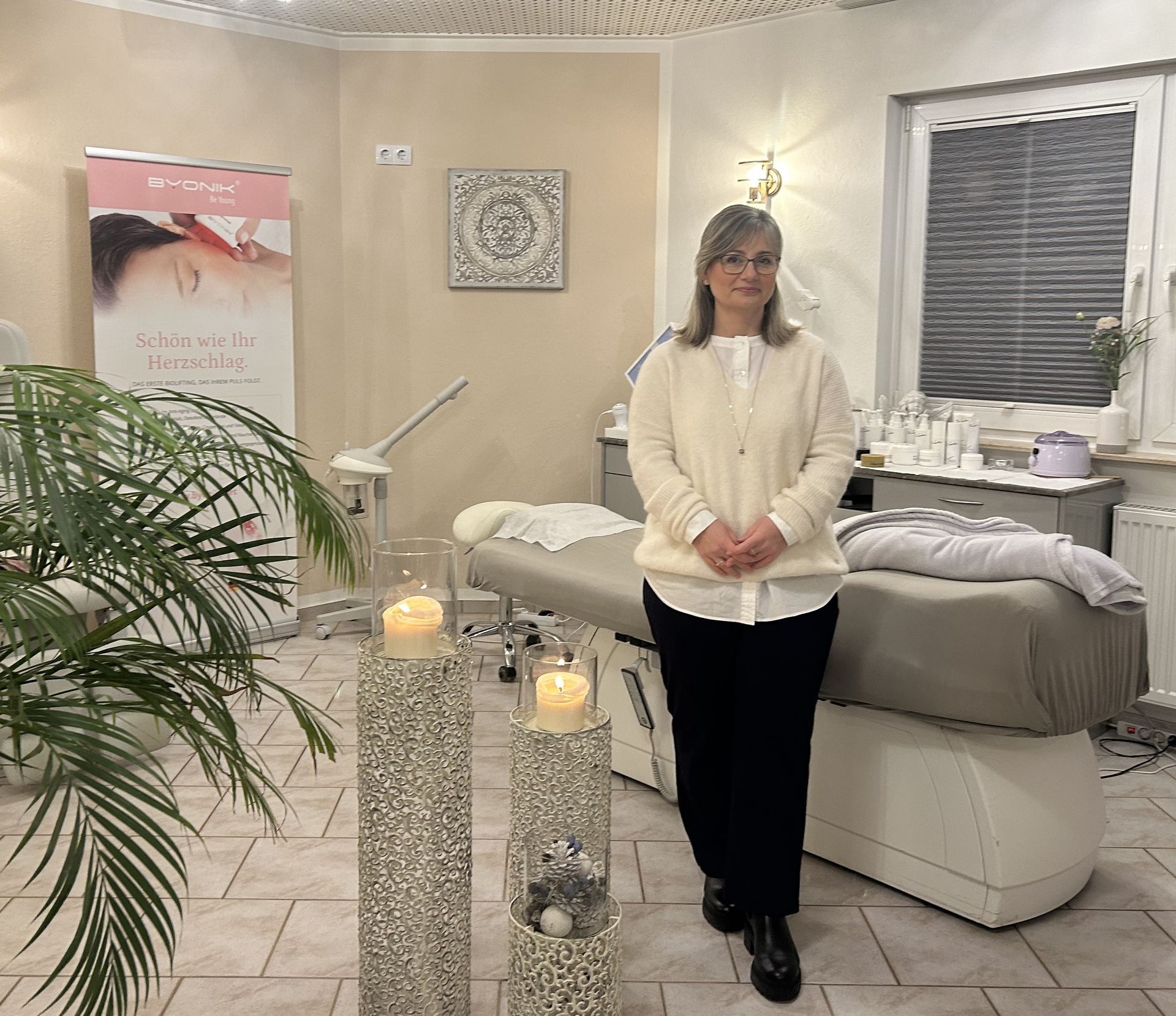 Ilka Schreiter Bild der Inhaberin Ilka Schreiter. Sie steht an einem Tresen, daneben sind Orchideen. Ein Engel und ein Sparschweinchen stehen auch auf dem Tresen.