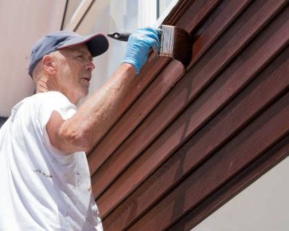 A man painting wood siding of a house with a paintbrush, focused on applying a fresh coat of paint.