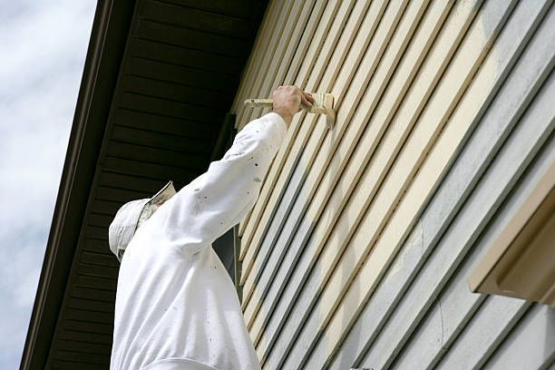 A man in white clothing is painting the exterior of a house with a paintbrush.