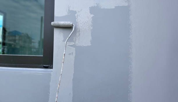 A person uses a paint roller to apply paint to the exterior wall of a house.