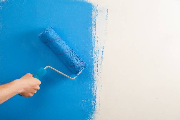 A person painting a blue wall in a home office using a roller, showcasing a fresh color choice for the space.