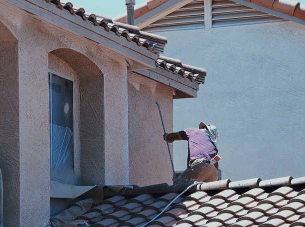 A man painting a roof on a house with wood siding focused on applying a fresh coat of paint.