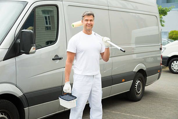 A man dressed in white pants and a shirt stands next to a van labeled