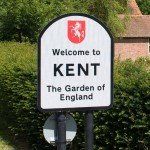 small sign with the words welcome to kent the garden of england on it with a small cottage behind