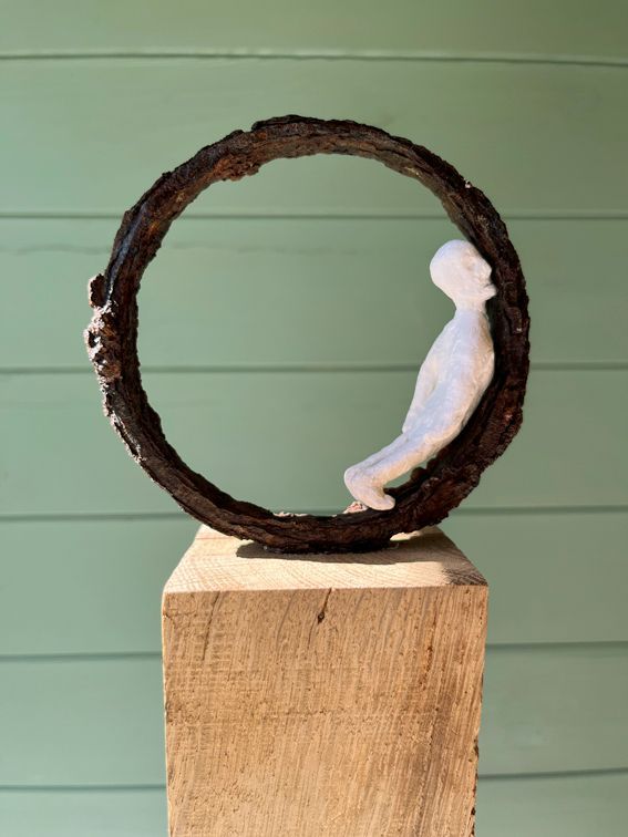 Skulptur aus Holzmaché anlehnend in einem rostigen Eisenring. Auf einem Holzklotz.