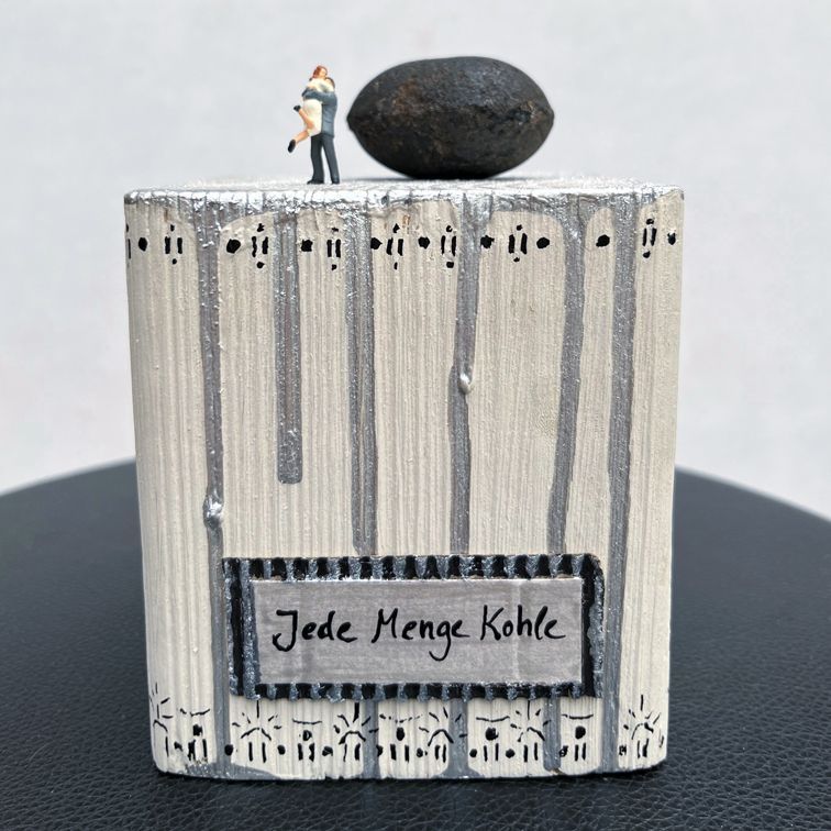 Bemaltes Holz mit Stück Eierkohle und einem Pärchen in Miniaturformat, dass sich in den Armen liegt.