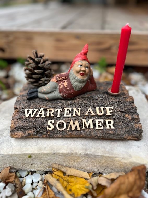 Antiker Gartenzwerg liegend auf Rinde mit Kerze und Tannenzapfen