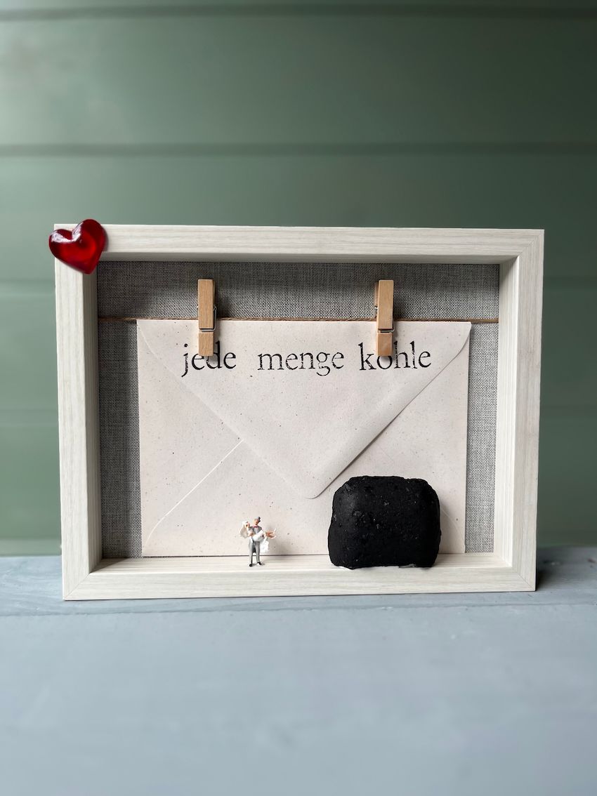 Briefumschlag an der Wäscheleine, Aufdruck jede Menge Kohle, darunter ein Hochzeitspaar im Miniaturformat und daneben ein Stück Holzkohle