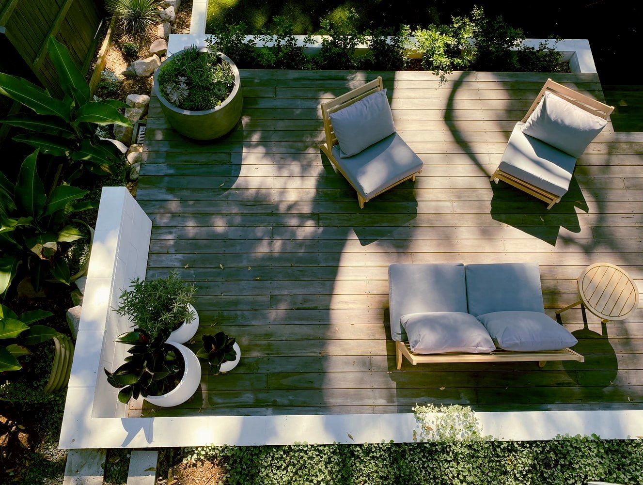 Terrasse aus Holz mit Betoneinfassung, darauf stehen grüne Pflanzen in weißen und grauen Blumentöpfe. Man sieht außerdem einen Teil des frischen Rasens