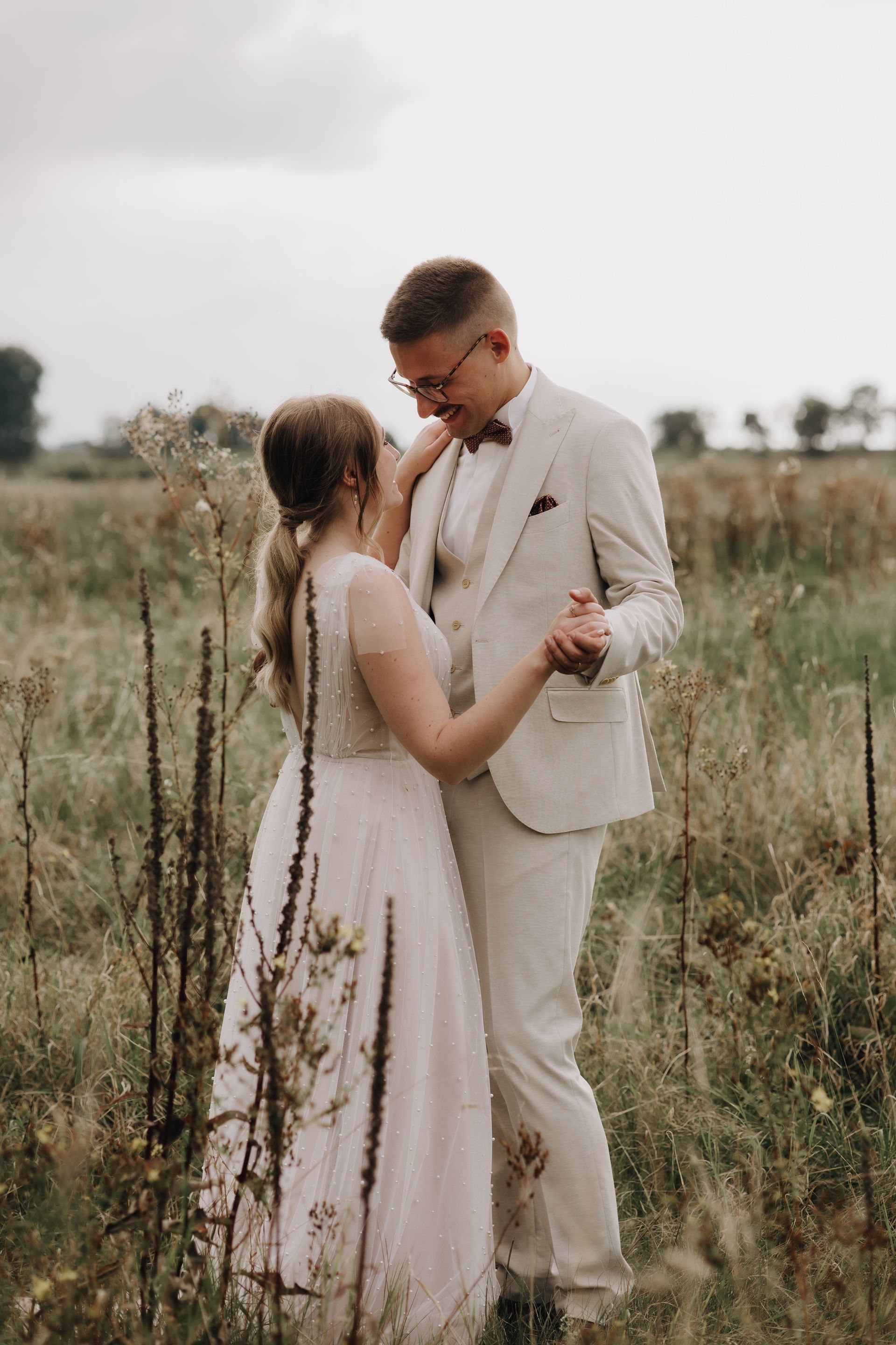 Brautpaar in einem Feld tanzend. Bräutigam in einem beigen Anzug im Bohostil und sie in einem Brautkleid mit Perlen im Bohostil. Hochzeitsfotografin in Köln. Haus Piwipp Dormagen
