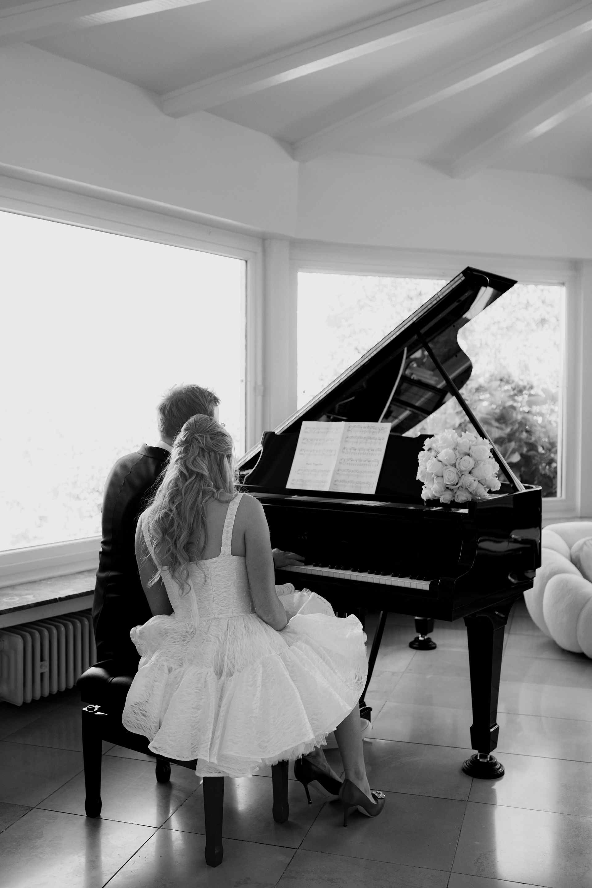 schwarz-weiß Fotografie von einem Hochzeitspaar an einem Klavier spielen. Die Braut ist anlehnend, es sieht aus wie ein Magazincover in einem kurzen Brautkleid mit Manolo Blano Schuhen in Marienheide bei Nümbrecht.
