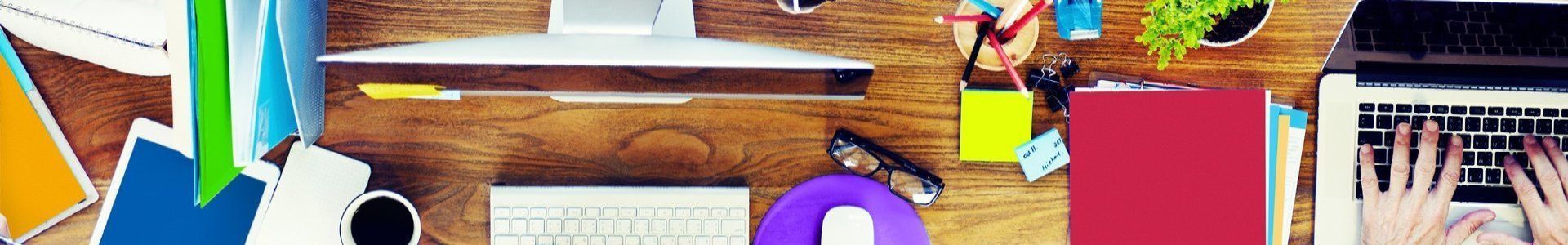 Schreibtisch von oben mit vielen Krimskrams darauf wie Brille, bunte Zettel, Klebezettel, Stifte, Stifthalter, bunte Hefte, Notizblöcke, PC Tastatur.