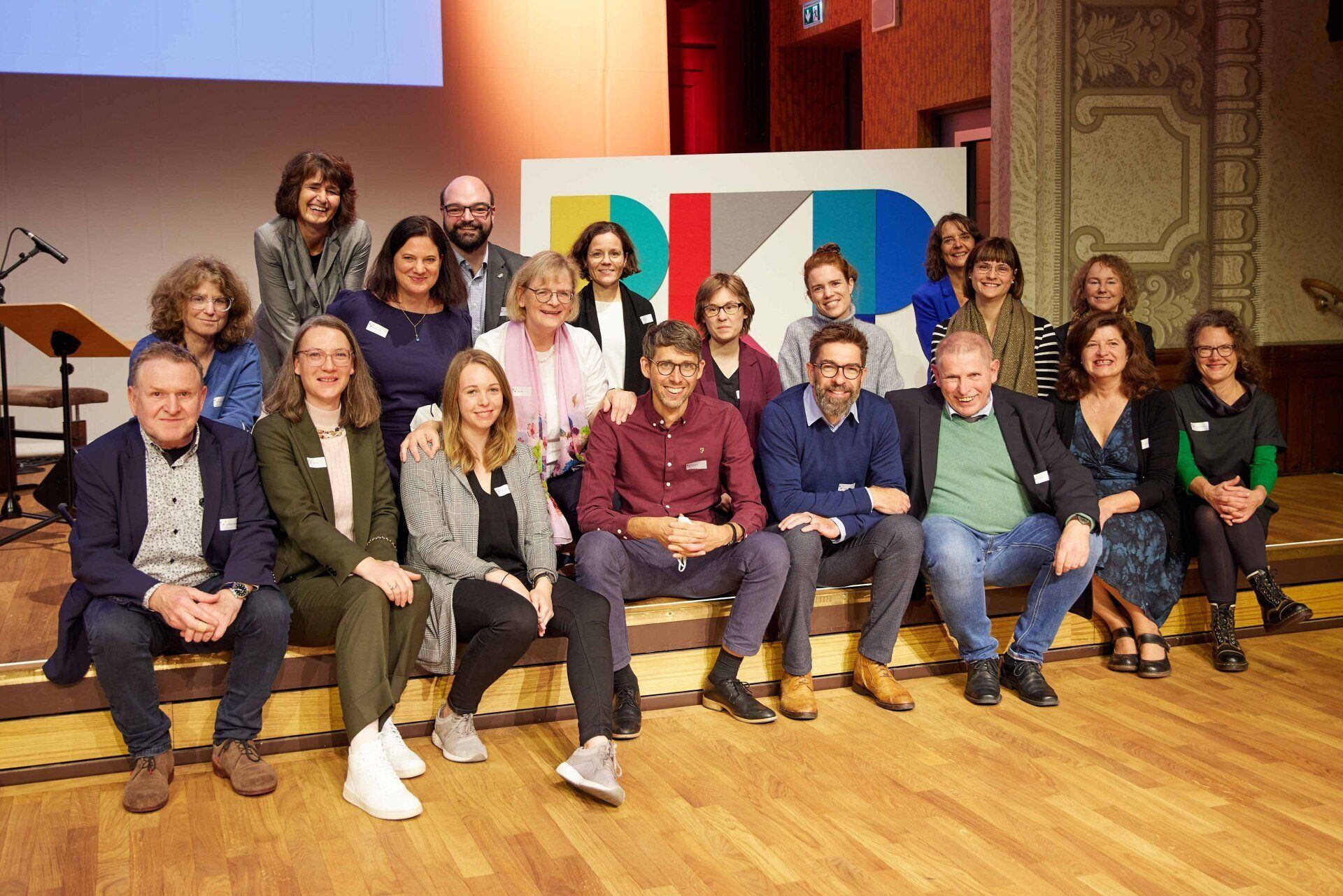 Gruppenbild aller RKP Büros. Die Mitarbeiter:innen sitzen und stehen auf der Bühne, dahinter das RKP-Logo.