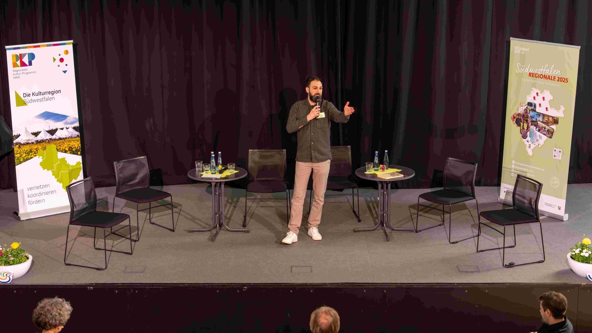 Murat Isboga steht auf der Bühne und gestikuliert. Rechts und links sind die Aufsteller der Kulturregion Südwestfalen und der Regionale Südwestfalen 2025 im Bild.