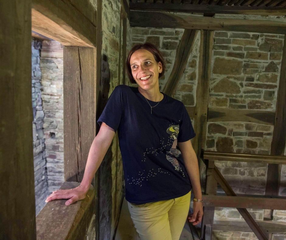 Barbara Peveling steht in einem Raum in der Wendener Hütte mit Fachwerk und Bruchsteinmauern. Sie lehnt sich seitlich an ein Fenster und schaut lächelnd hinaus.