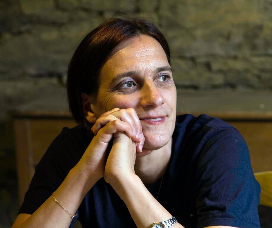 Die Autorin sitzt in der Wendener Hütte an eineme Tisch, die Ellebogen aufgestützt und das Gesichts in die gefalteten Hände seitlich abgelegt.