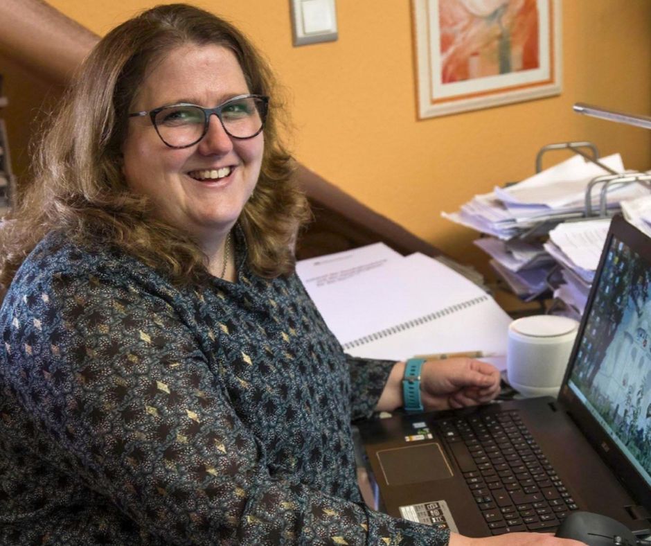 Bettina Görlitzer, die an ihrem Schreibtisch vor ihrem aufgeklappten Laptop sitzt und lacht. Der Schreibtisch ist überfüllt mit Zetteln, Arbeitspapieren, Notizblöcken.