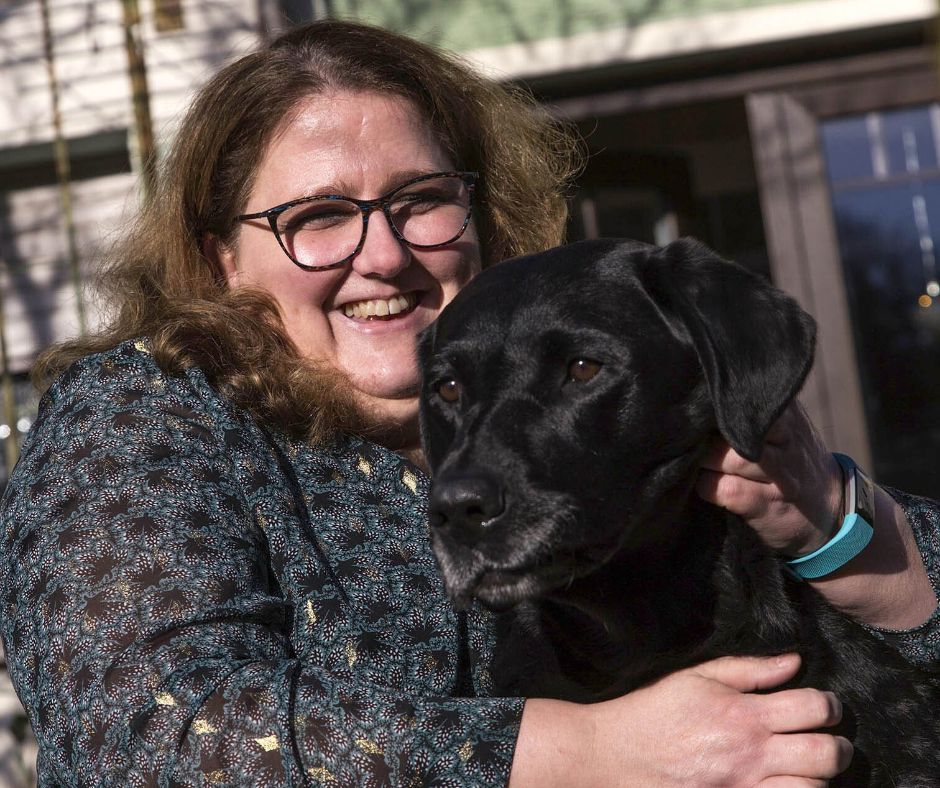 Porträt von Bettina Görlitzer mit Hund, einem kräftigen schwarzen Labrador, der von ihr gekrault wird.