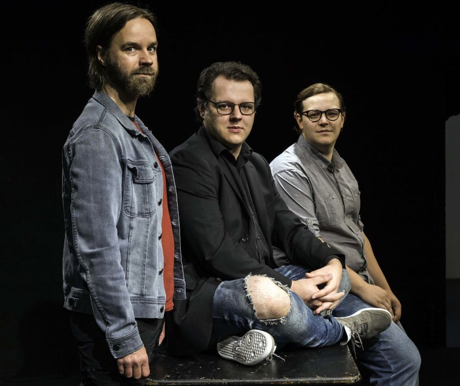 David sitzt in der Mitte eines abgestoßenen schwarzen Tisches, links neben steht Milan, rechts lehnt sich Tim an den Tisch an. Alle drei tragen legere Kleidung, der intergrund ist ebenfalls schwarz.