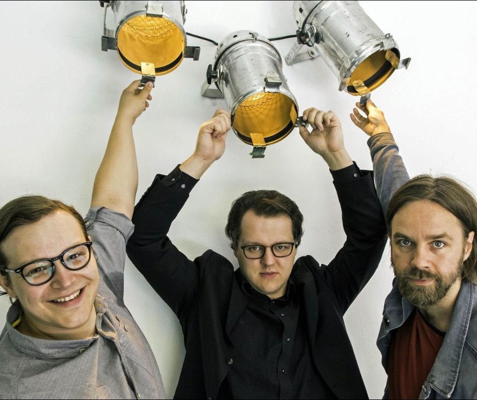 Tim, David und Milan richten je der drei einen Scheinwerfer, die sich über ihren Köpfen befinden, auf einen fiktiven Besucher und schauen auffordernd in die Kamera.