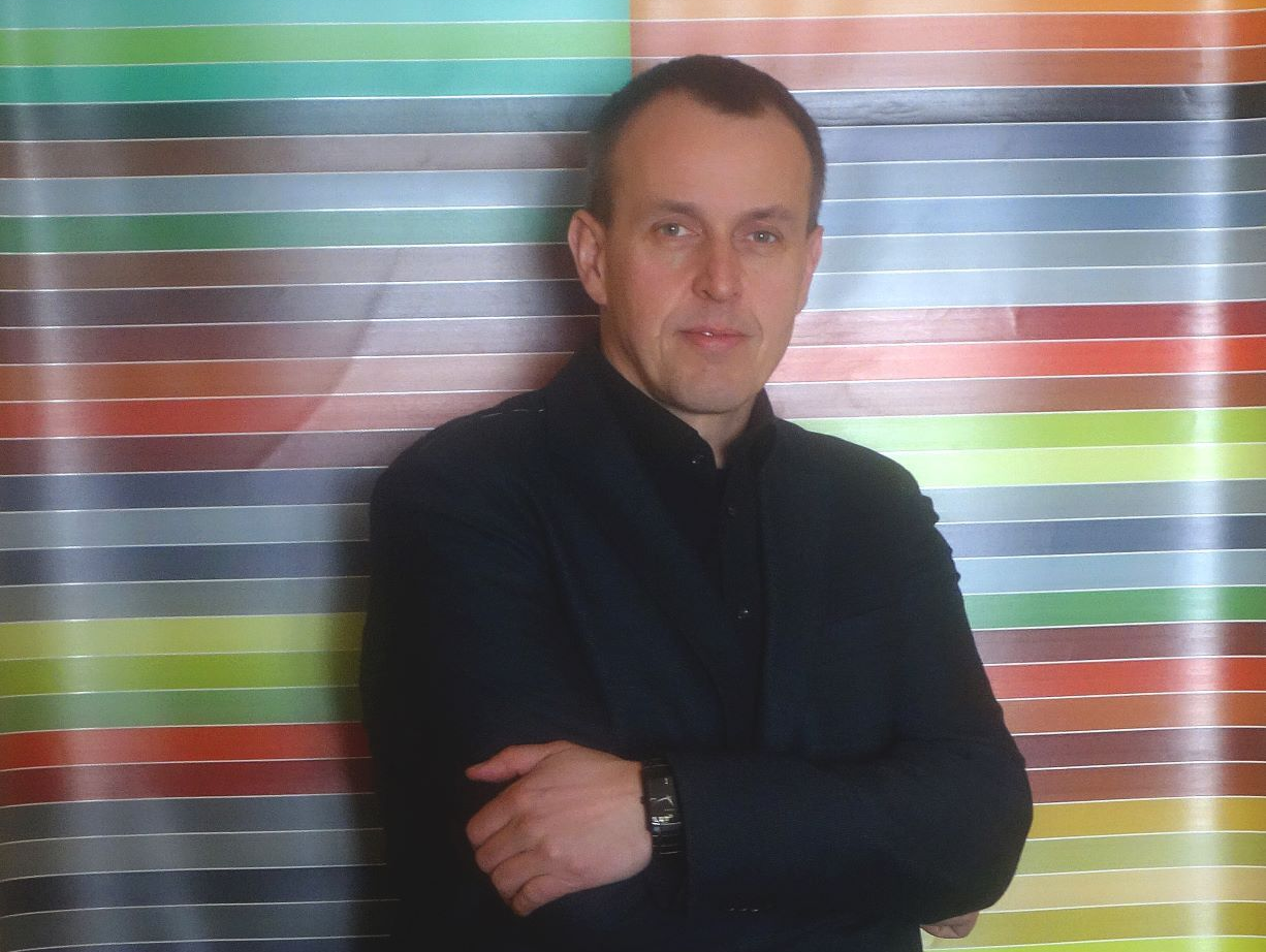Herr Krüger steht vor einem bunten Plakat und blickt in die Kamera