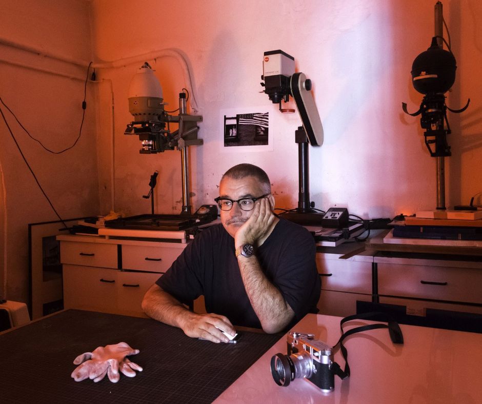 Dirk Vogel sitzt in seinem Fotolabor im Keller an einem Tisch, den Kopf in die Hand gestütz schaut er sinnierend vor sich hin. Im Hintergrund sieht man die Geräte zur Filmentwicklung, auf dem Tisch neben ihm liegen Kamera und weiße Handschuhe.
