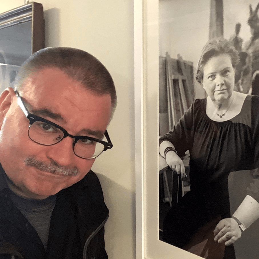 Dirk Vogel schaut seitlich in die Kamera, rechts  neben ihm hängt ein schwarz-weiß Foto einer Künstlerin, das er fotografiert hat.