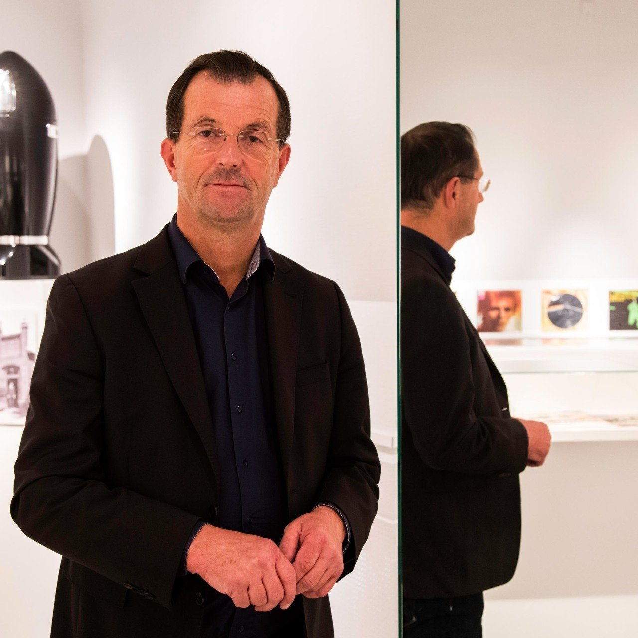 Dr. Trox steht in einem Ausstellungraum, im Hintergrund sind Exponate zu sehen. Links neben ihm steht ein Spiegel, der die Rückansicht des Museumsleiters inklusive weitere Exponate spiegelt.