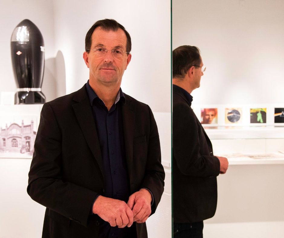 Dr. Trox steht in einem Ausstellungraum, im Hintergrund sind Exponate zu sehen. Links neben ihm steht ein Spiegel, der die Rückansicht des Museumsleiters inklusive weitere Exponate spiegelt.