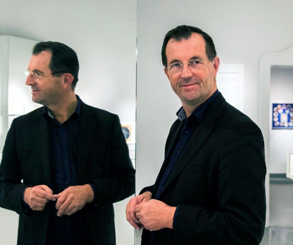 Der Museusleiter seht in einem Ausstellungsraum, im Hintergrund ist eine Vitrine und ein Bild zu sehen. Er steht vor einem Spiegel und schaut in die Kamera, gespiegelt wird das seitliche Profil.