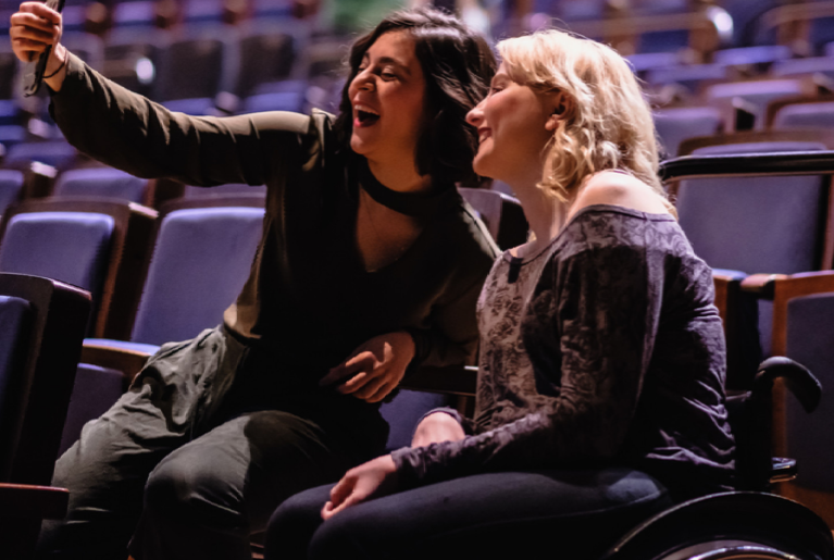 Zwei Frauen sitzen nebeneinander im Theater. Sie lachen in die Handykamera, die eine der beiden Frauen in der Hand hält. Eine der beiden Frauen sitzt im Rollstuhl.