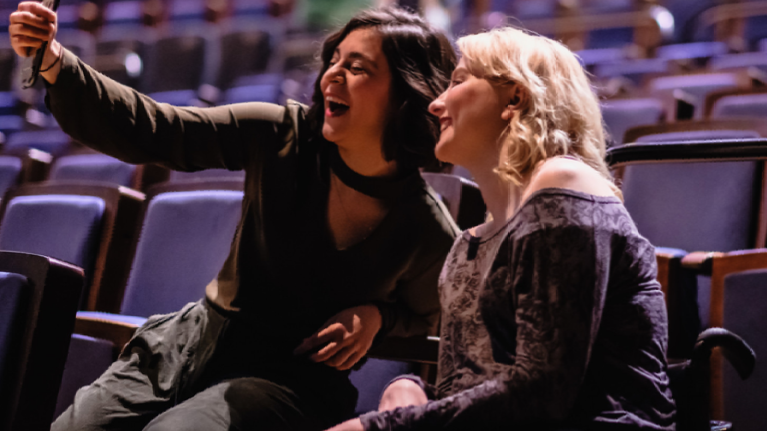 Zwei Frauen sitzen nebeneinander im Theater. Sie lachen in die Handykamera, die eine der beiden Frauen in der Hand hält. Eine der beiden Frauen sitzt im Rollstuhl.