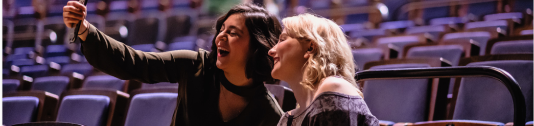 Zwei Frauen sitzen nebeneinander im Theater. Sie lachen in die Handykamera, die eine der beiden Frauen in der Hand hält. Eine der beiden Frauen sitzt im Rollstuhl.