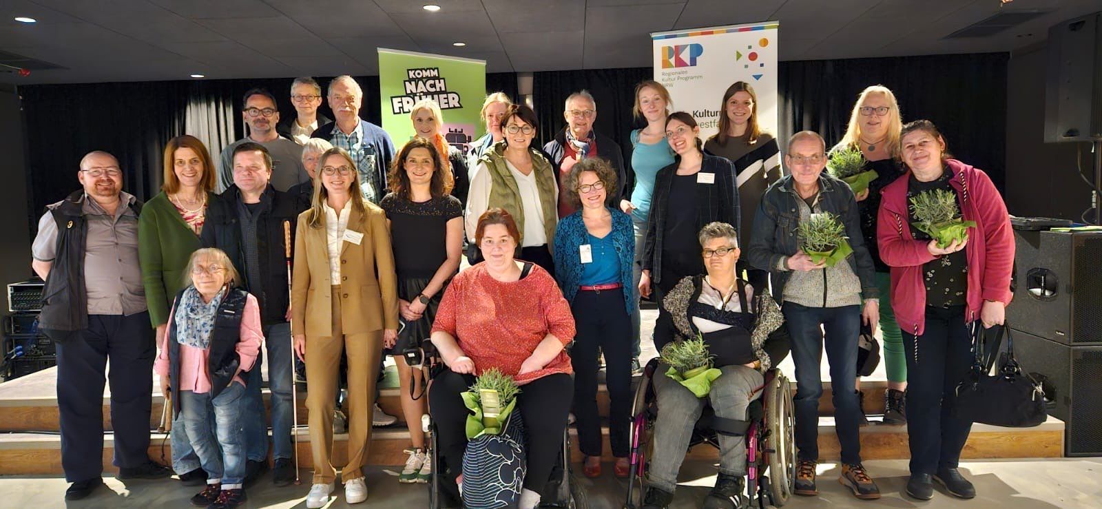 Gruppenfoto der Teilnehmenden des Praxisworkshops. Ungefähr 25 Menschen mit und ohne Behinderung stehen zusammen auf und vor der kleinen Bühnen der OT Grevenbrück.