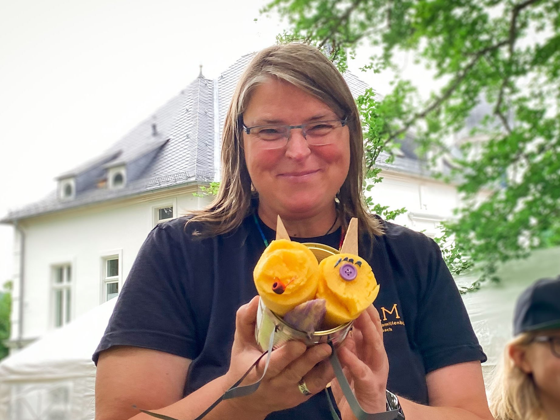 Frau Kühn steht in einem Park und lächelt in die Kamera. In der Hand hält sie eine Dose aus der ein aus Gemüse gebasteltes Tier schaut