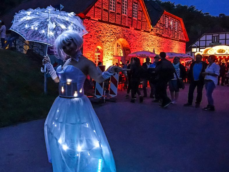 Eine Dame in weißem durchsichtigen Reifrockkleid und Schirm, beides mit LEDs beleuchtet steht vor der rot beleuchteten Luisenhütte. Es dämmert, im Hintergrund ist eine Menschenmenge.