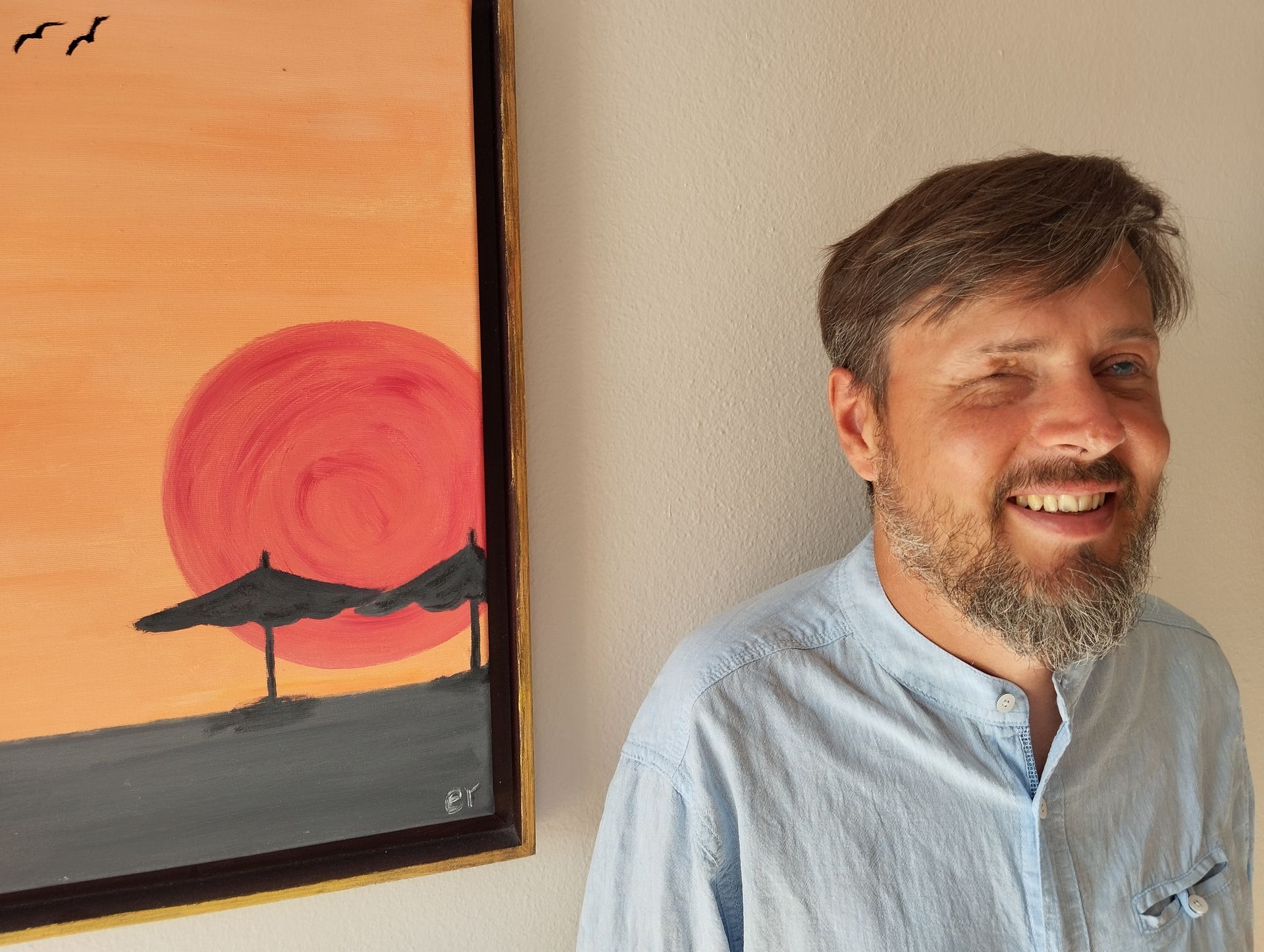 Jan Meyer-Krügel, blind, steht vor einem Gemälde, auf dem ein Strand und eine glühende Sonne abgebildet  sind. Er trägt kurze Haare, einen Bart und ein Baumwollhemd.