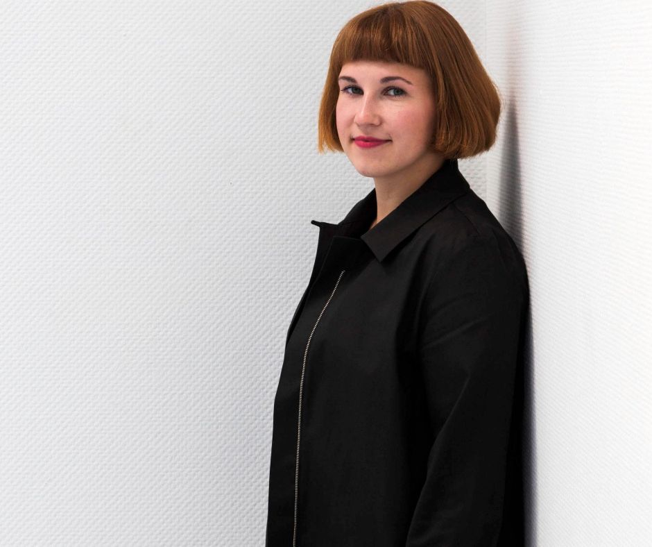 Jennifer Cierlitza, schwarz gekleidet, steht in einer schlichen weißen Zimmerecke und blickt in die Kamera.