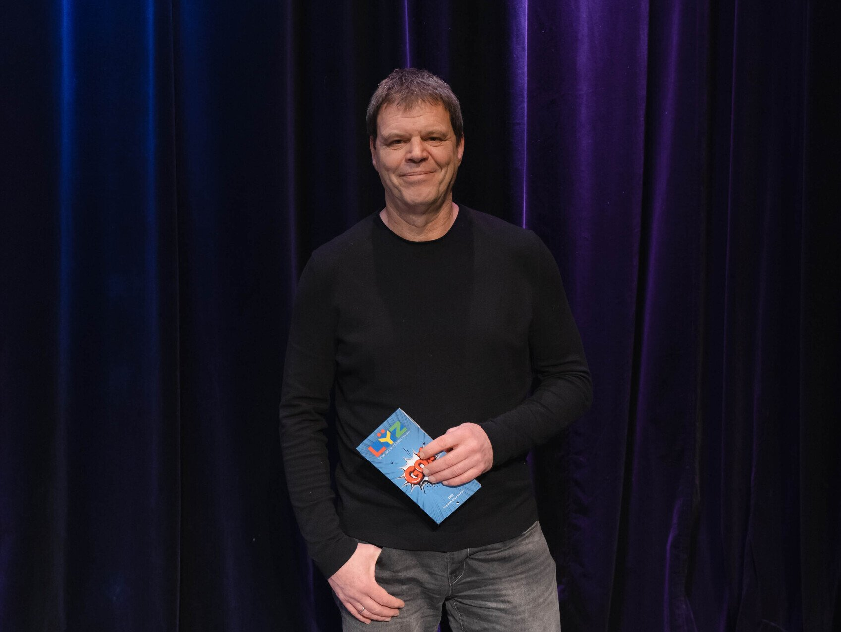 Herr von Heyden steht vor einem blauen Theatervorhang und schaut in die Kamera